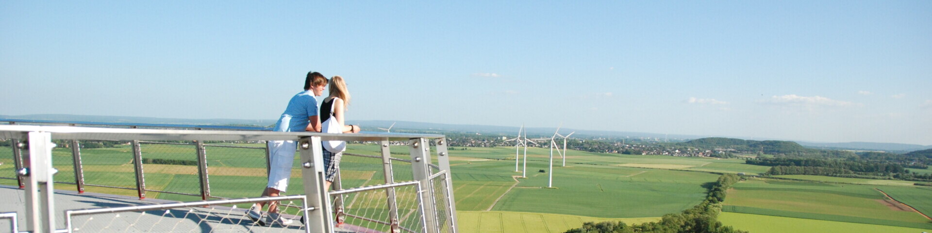 Zwei Menschen stehen auf einer Aussichtsplattform. Im Hintergrund sind eine grüne Landschaft und Windräder zu sehen