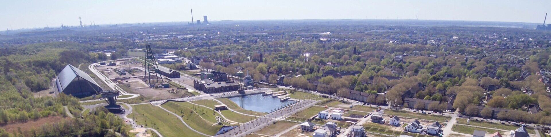 Ein Park mit viel Grün und ein paar kleinen Gebäuden. In der Mitte ein künstlicher See, im Hintergrund sehr viele Bäume