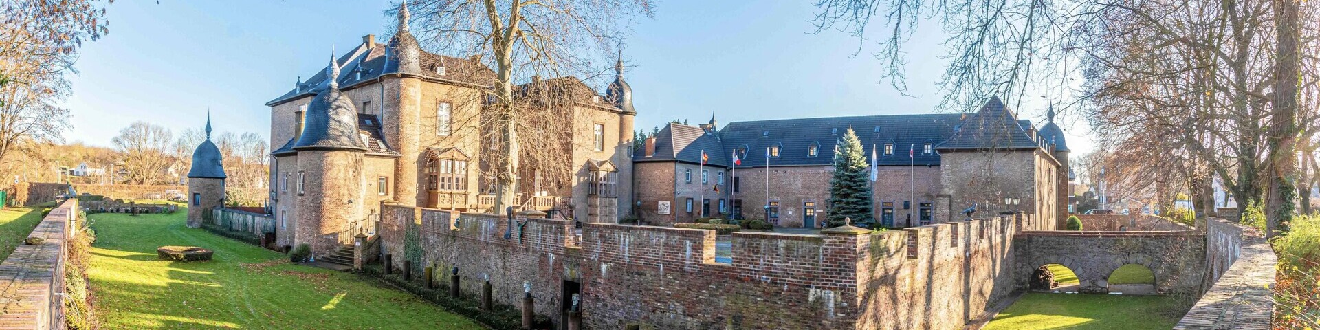 Ein historisches Gebäude mit einer Steinmauer ringsherum und einem Burggraben mit Rasen
